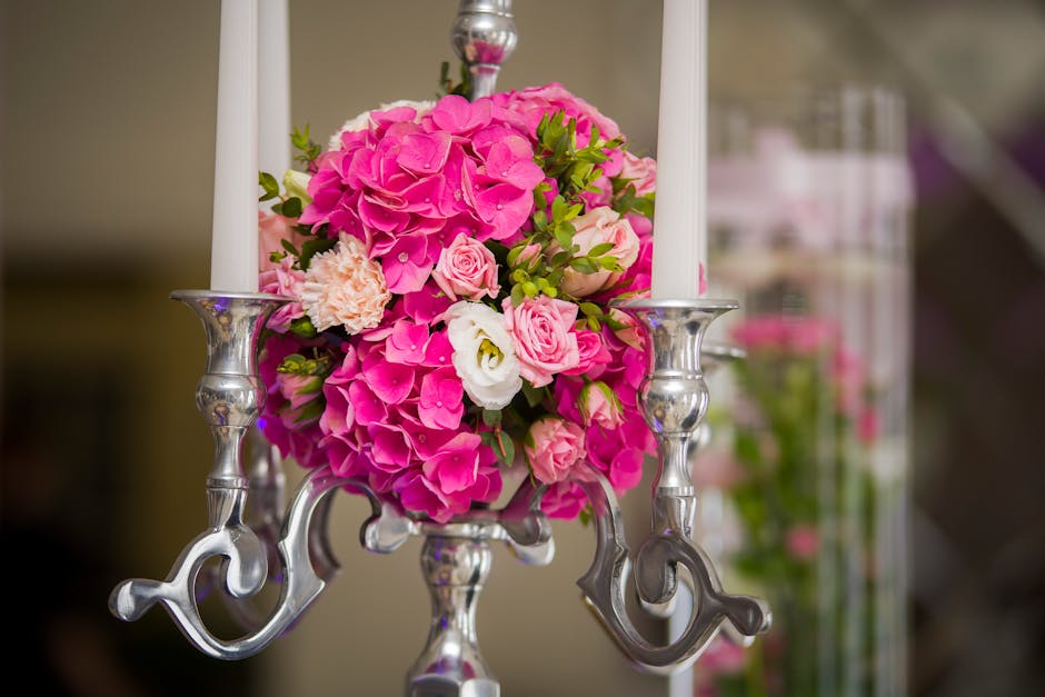 A floral arrangement featuring a vibrant bouquet of pink hydrangeas, soft pink roses, and white lisianthus, accented with lush green foliage. The bouquet is placed in a metallic silver candelabrum wit