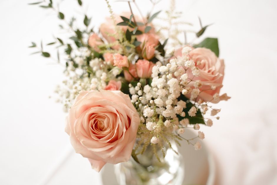 A bouquet arrangement featuring soft pink roses, complemented by white baby's breath and sprigs of greenery, all arranged in a clear glass vase. The roses have full, layered petals with a delicate, ve