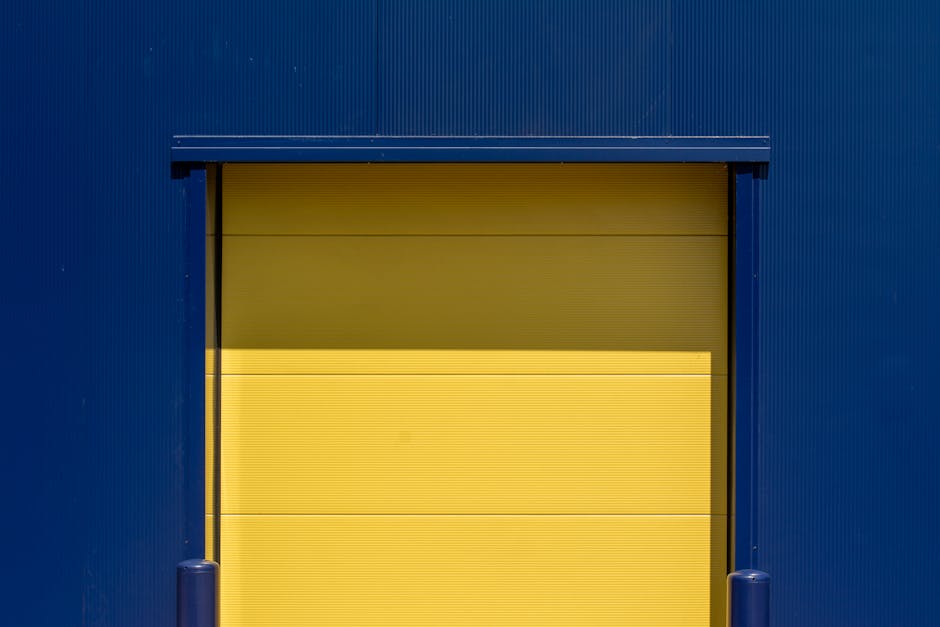 A closed loading bay door set into a blue industrial building wall, with a central yellow rolling shutter. The surrounding blue structure is ribbed metal siding, and there are two blue bollards at the