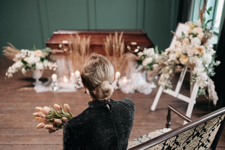 A woman with blonde hair styled in a low bun, dressed in a black velvet top, is seated on a vintage-patterned bench and holding a bouquet of pink tulips. She is observing a funeral floral arrangement