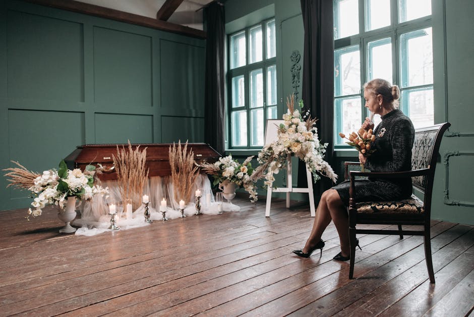 An elegant woman dressed in a black lace dress and black heels is seated on an ornate wooden chair inside a softly lit room with green paneled walls and large windows. She holds a small, delicate flor