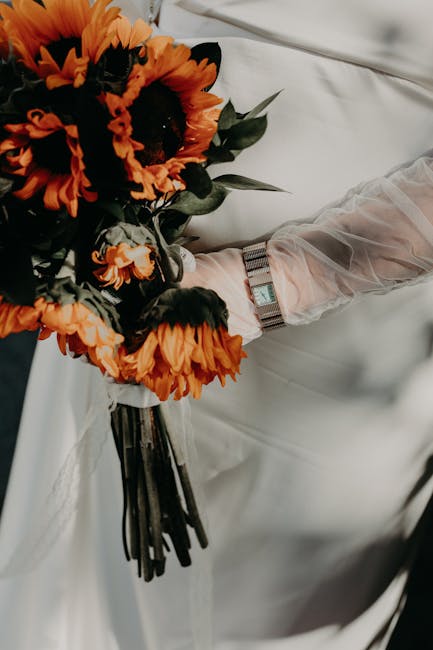 A close-up of a bouquet of fresh orange and black-eyed sunflower flowers with vibrant petals and dark centers, wrapped in white paper and tied with a dark ribbon, being held by a person wearing a shee