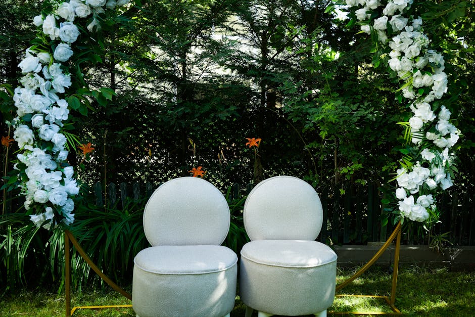 A floral ceremony arch featuring cascading white roses and green foliage frames two elegant white upholstered chairs with rounded backs and cushioned seats, set against a lush garden background with d
