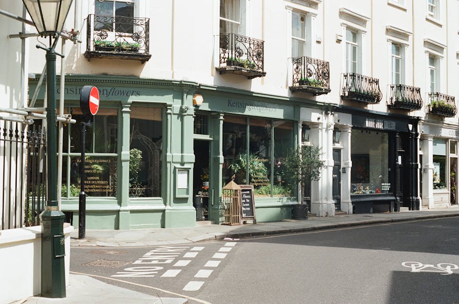 A storefront with large glass windows displaying various floral arrangements and greenery, painted in a soft pastel green color, located on a street corner. The shop has a sign that reads 'Kensington