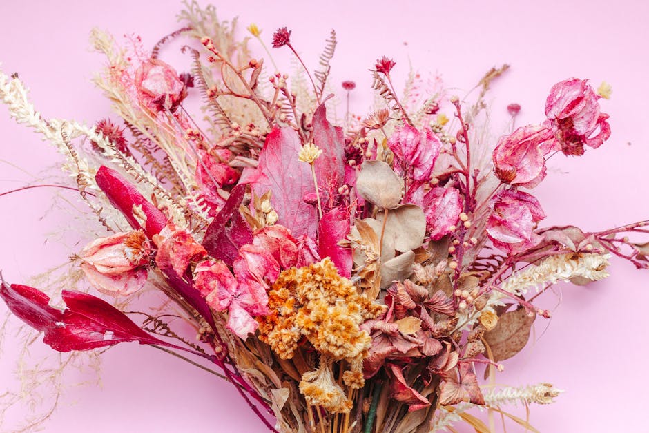 A bouquet of dried flowers arranged on a soft pink background, featuring various textures and shades of pink, cream, and beige. The bouquet includes dried roses, lilies, and small accent flowers, wrap